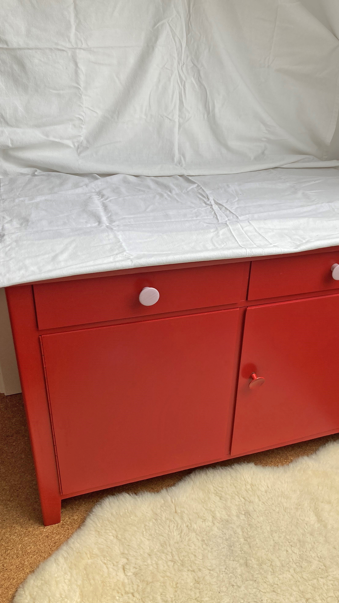 A red chest of drawers is covered in fabric with a lambskin in front of it.
